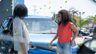 Two people smile and chat beside a blue car as confetti falls around them.